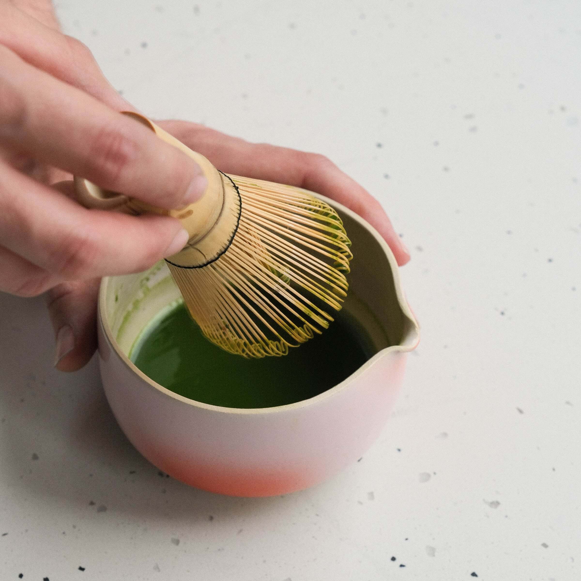 Hand holding a bamboo matcha whisk in a bowl mixing vibrant green matcha tea.