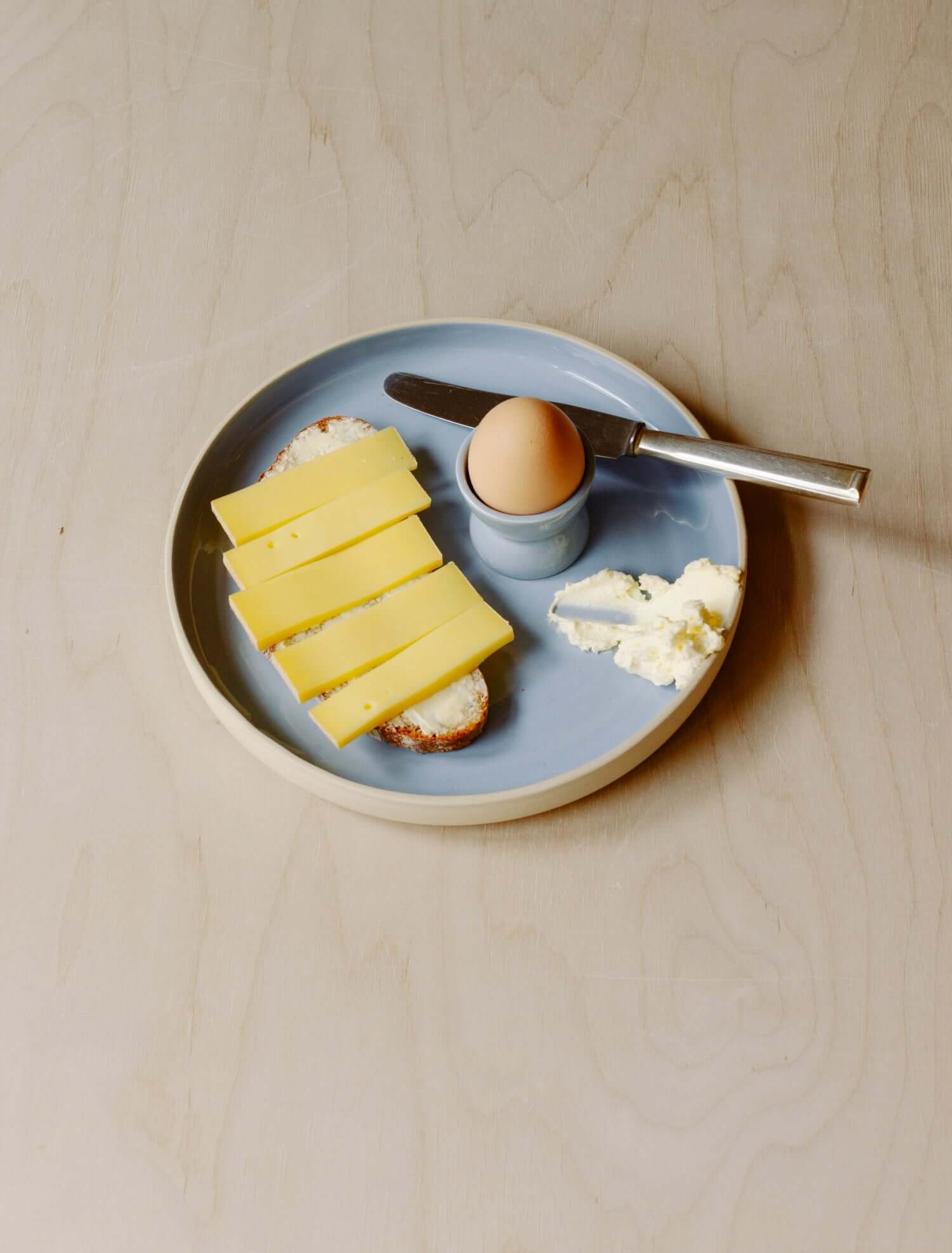 Plate with sliced cheese, an egg in an egg cup, and butter on a wooden table.