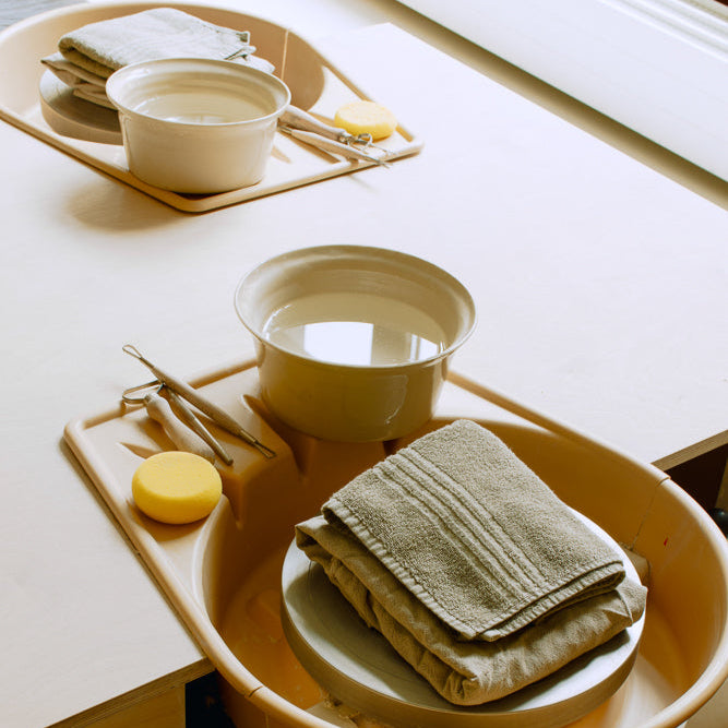 Pottery tools, water bowls, and towels set up for a beginner wheel class on a workshop table.