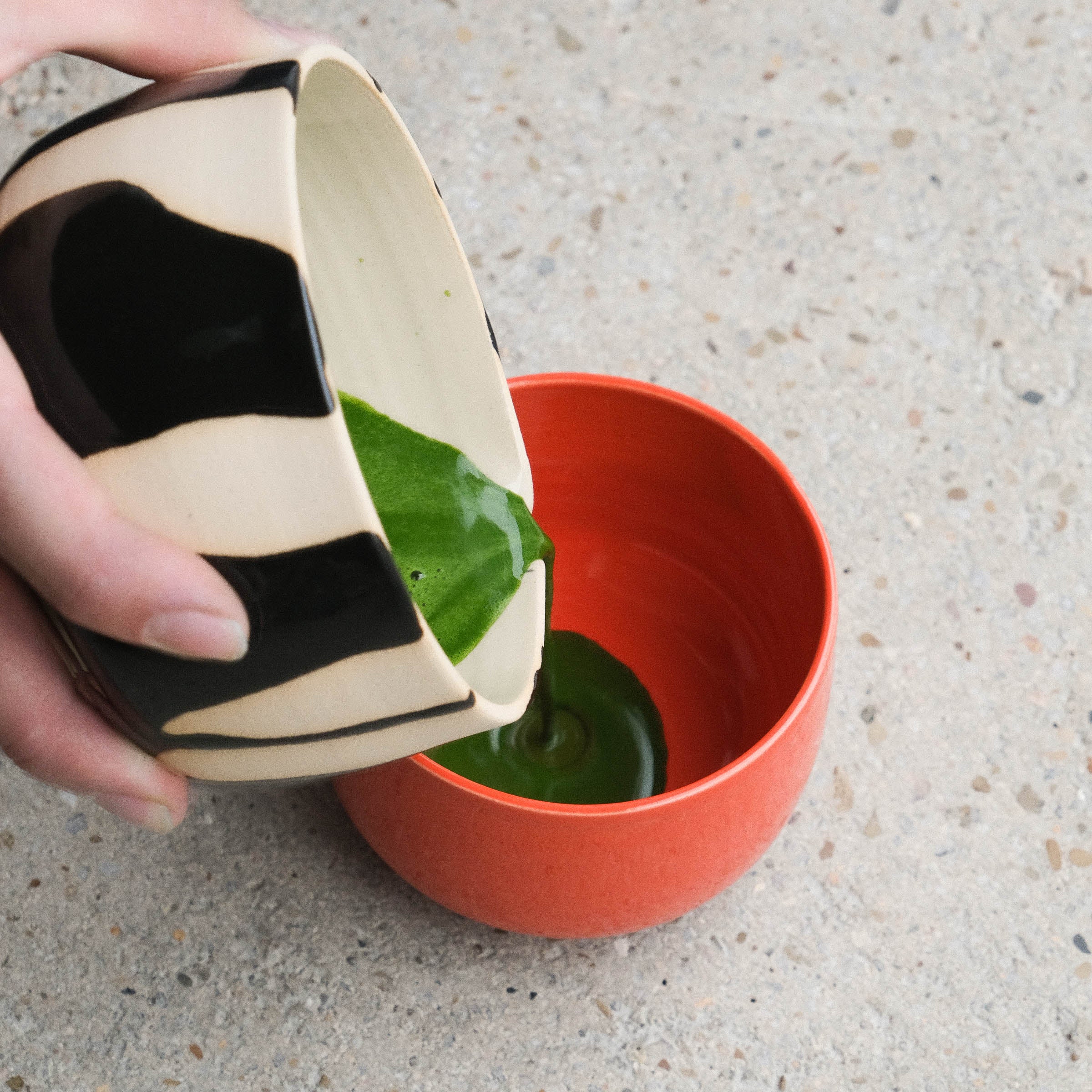 Pouring matcha from a unique serving bowl into a glossy red matcha cup made of stoneware.