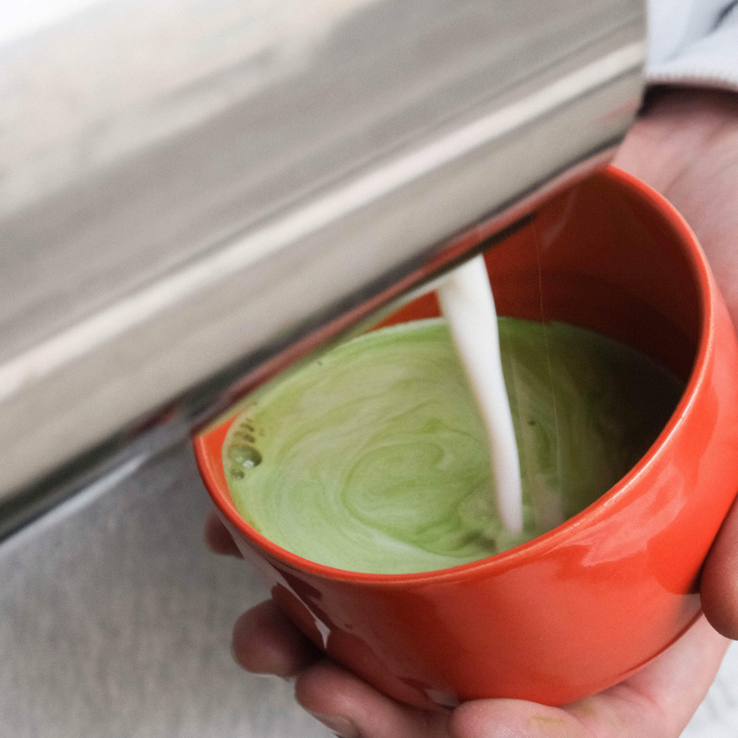 Pouring steamed milk into a glossy red matcha cup, creating a creamy green matcha drink.