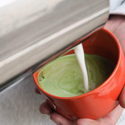 Pouring steamed milk into a glossy red matcha cup, creating a creamy green matcha drink.